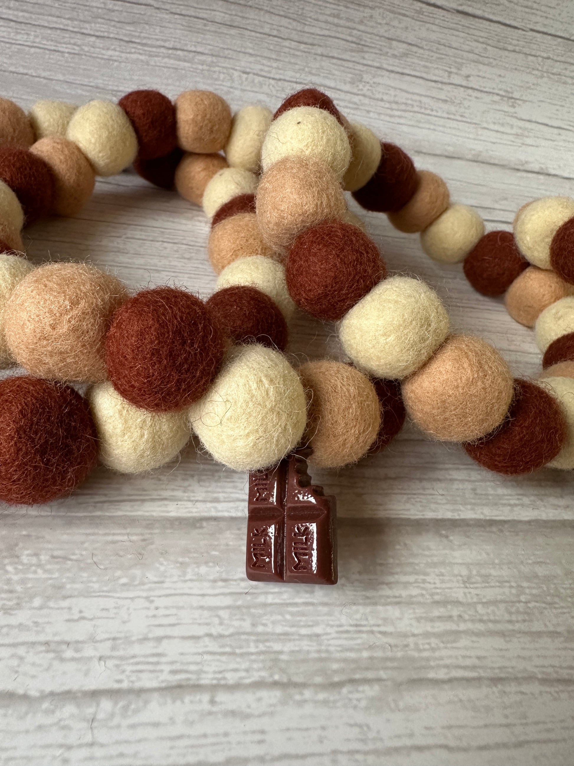 A close-up of Crafts by Kate's PomPom Pet Collar - Chocolate Lover features wool felt balls in cream, beige, and brown with a brown metal cross charm on a wooden surface.