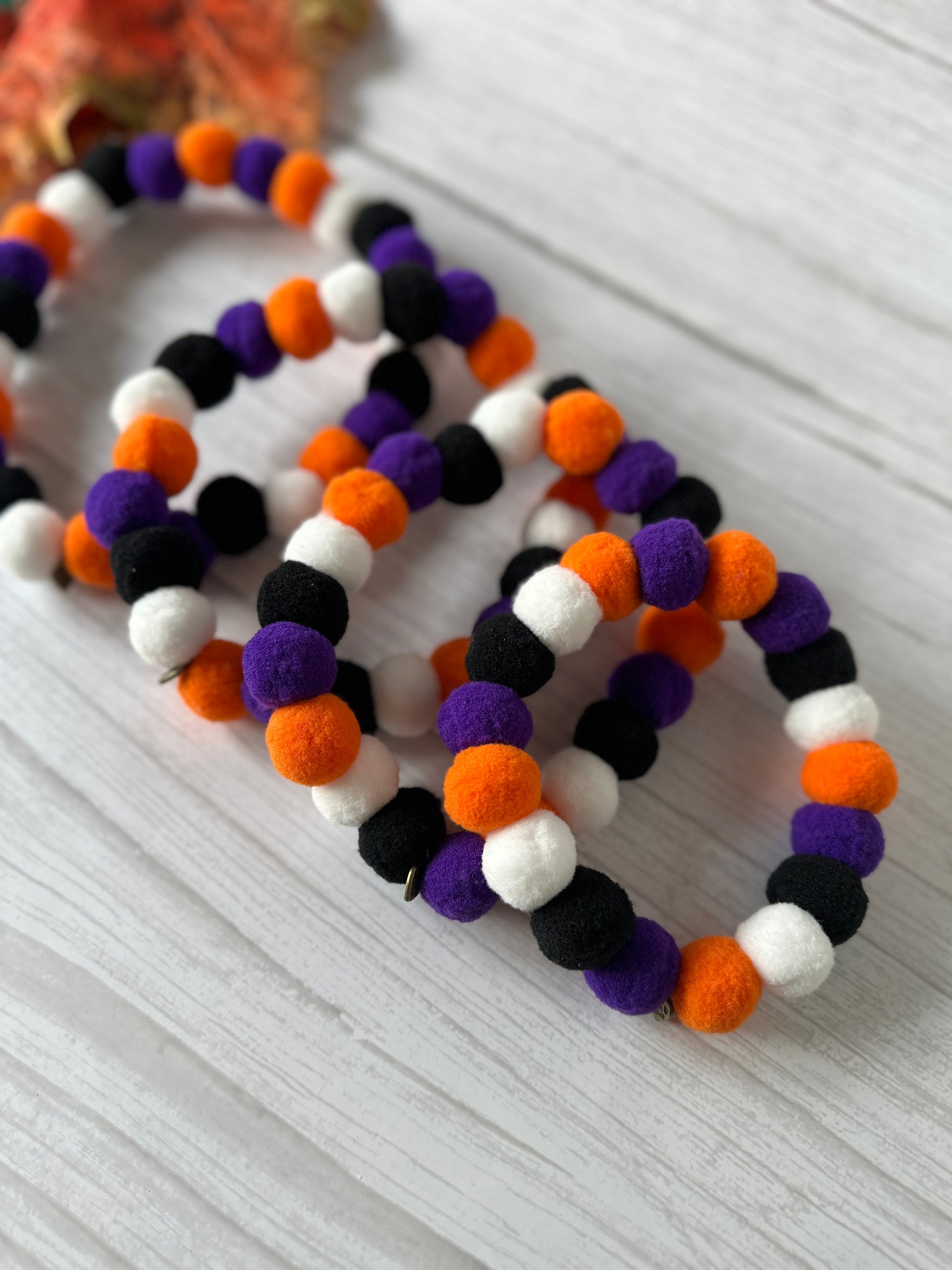 Crafts by Kate's PomPom Pet Collars - Halloween, featuring colorful elasticated bands adorned with small, round, fuzzy balls in shades of orange, purple, black, and white, are laid out on a light-colored wooden surface. The collars are arranged side by side for a playful and festive look perfect for the Halloween season.