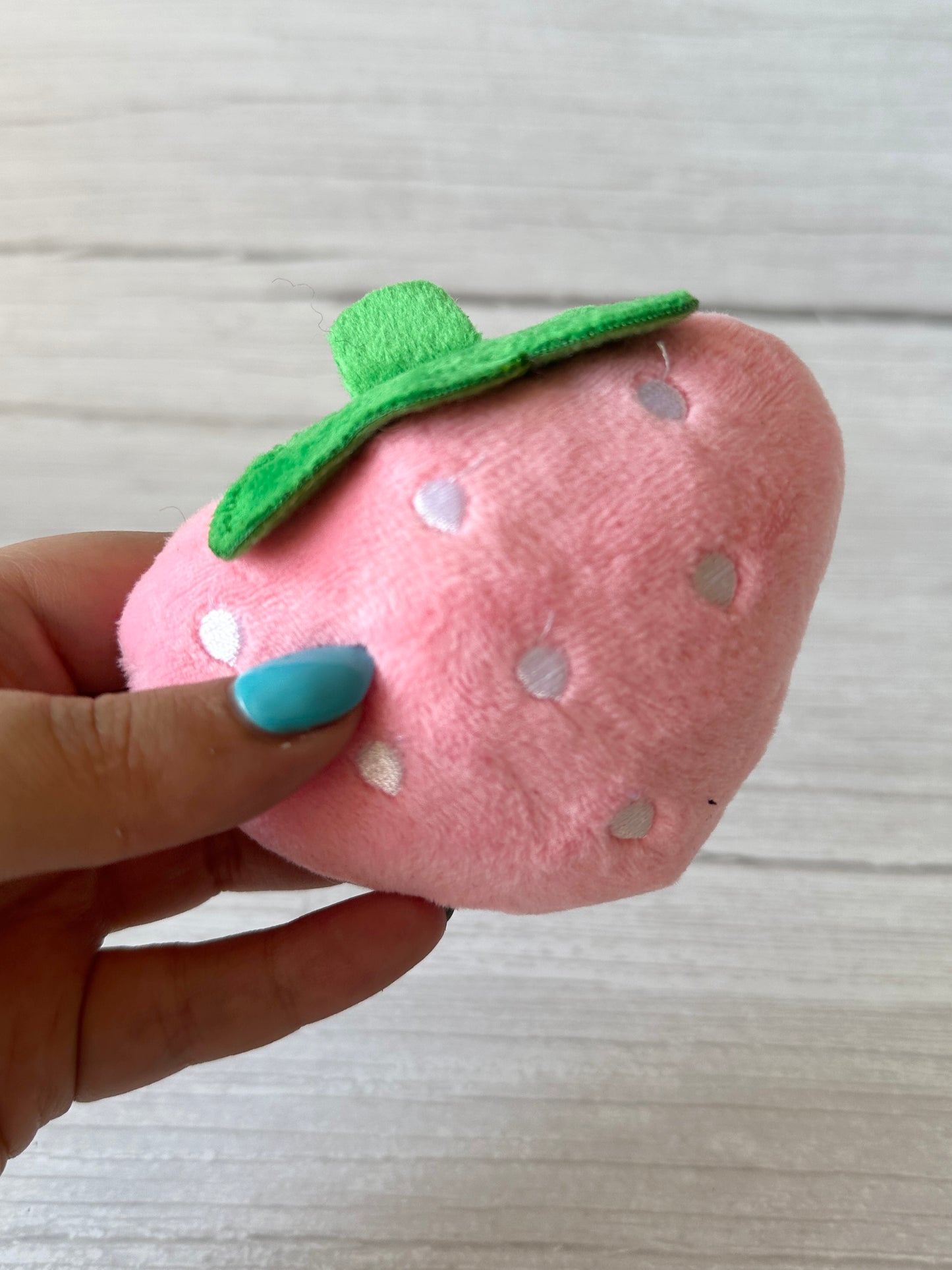 A hand with teal nail polish holds the Strawberries & Daisies - Arnie & Mac’s Goodies pet toy by Crafts by Kate— a soft, plush pink strawberry with white spots and green felt leaf—against a light wooden background.
