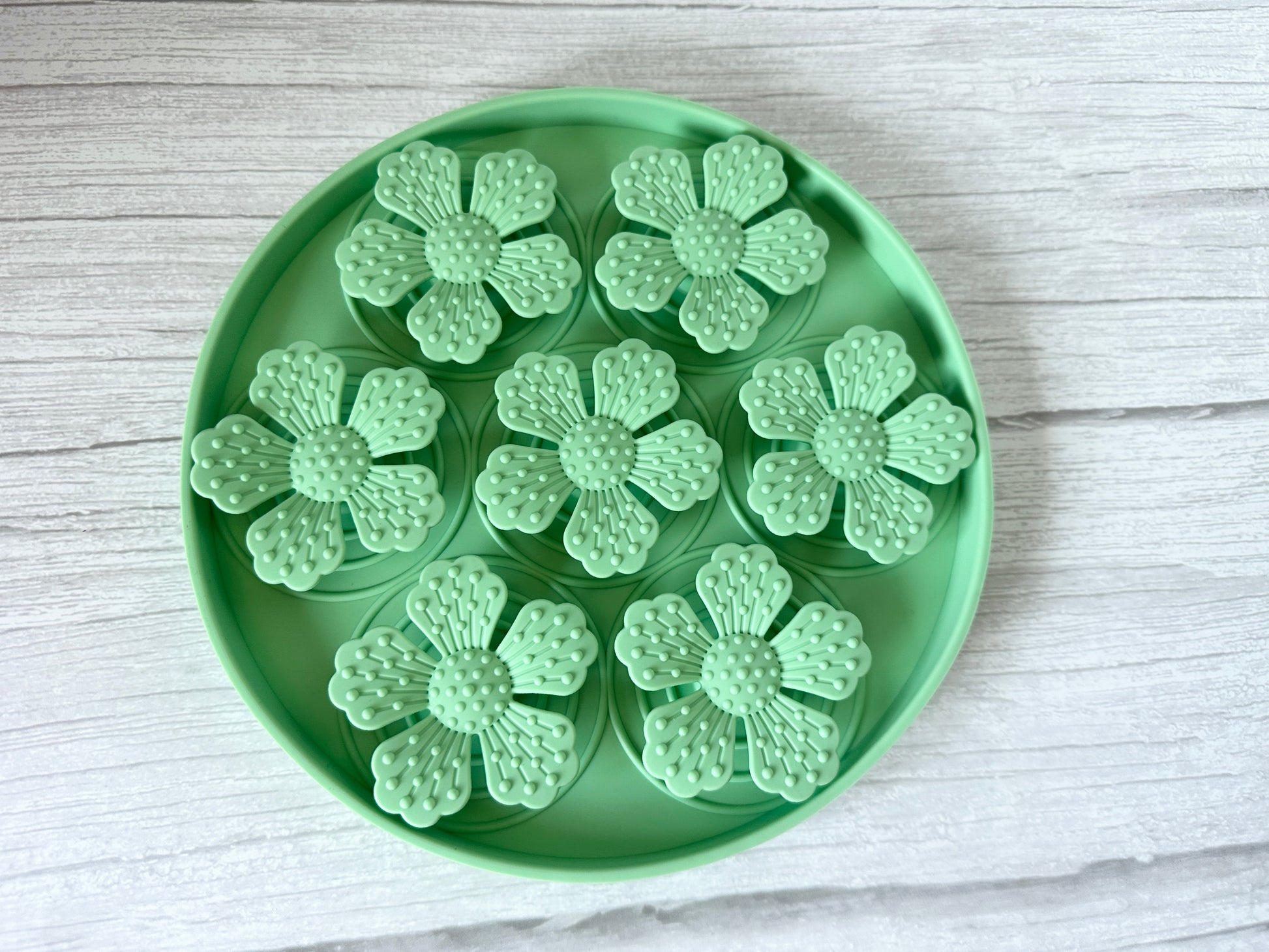 The round green Slow Feeder Pet Mat by Crafts by Kate features suction cups and holds seven mint-green, flower-shaped plastic items with detailed petals and textured centers. The tray sits securely on a light wood surface.