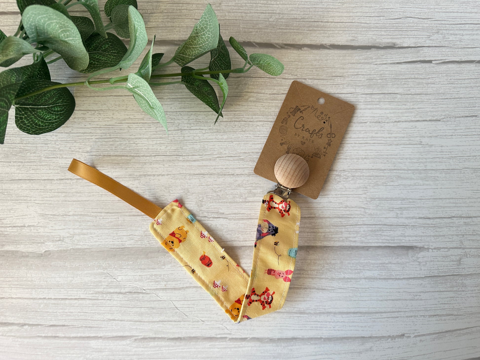 A Winnie the Pooh-themed yellow fabric pacifier clip made from ultra-soft cotton, featuring colorful animal patterns by Crafts by Kate, lies on light wood with a sprig of green leaves in the top left corner.