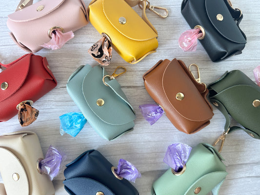 A collection of colorful Pet Boo Bag Holder Handbags by Crafts by Kate is displayed on a wooden surface. Each small faux leather pouch features a roll of plastic bags and a metal clip, available in red, pink, yellow, green, navy, and beige.