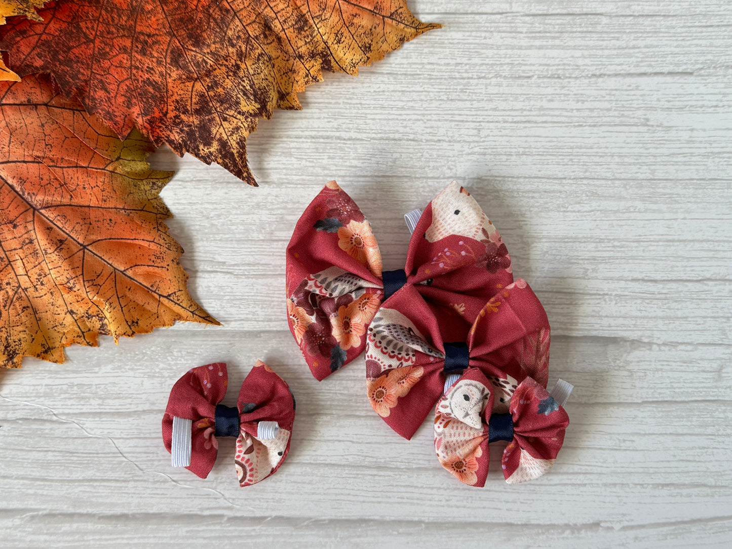 Three beautifully crafted woodland animals fabric bows by Crafts by Kate, showcasing charming floral patterns in maroon, are elegantly arranged on a white wooden surface. These cotton pet bows with collar attachments lie gracefully, enhanced by two large orange autumn leaves in the background for a touch of seasonal warmth.