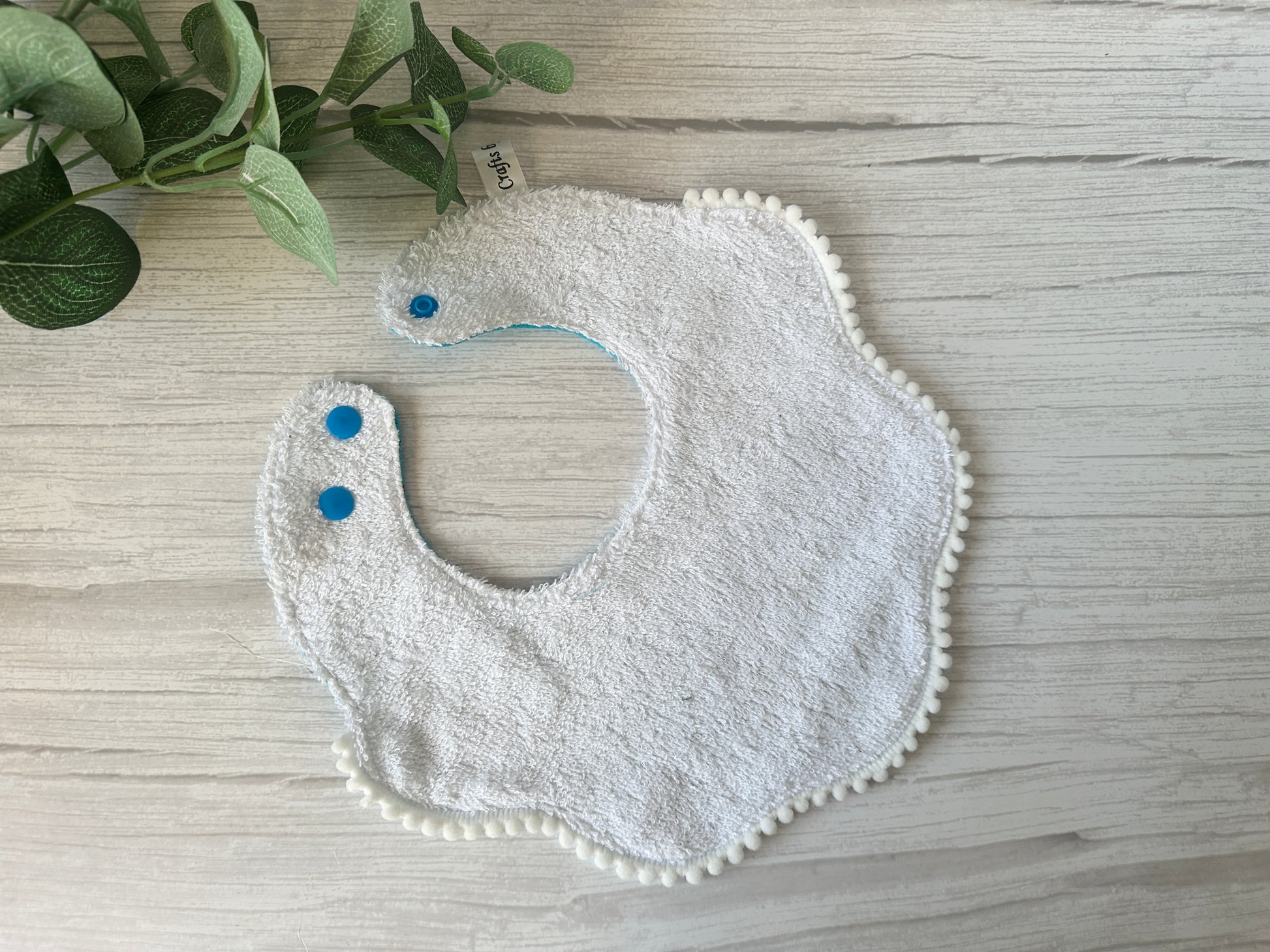 A crafted "Gruffalo in blue" baby bib from Crafts by Kate's Cotton & Terry Towelling Gift Set, with scalloped edges, blue snap buttons, and pom-pom trim, lies on a light wood surface. A sprig of green leaves rests in the top left corner. All made from ultra-soft cotton.