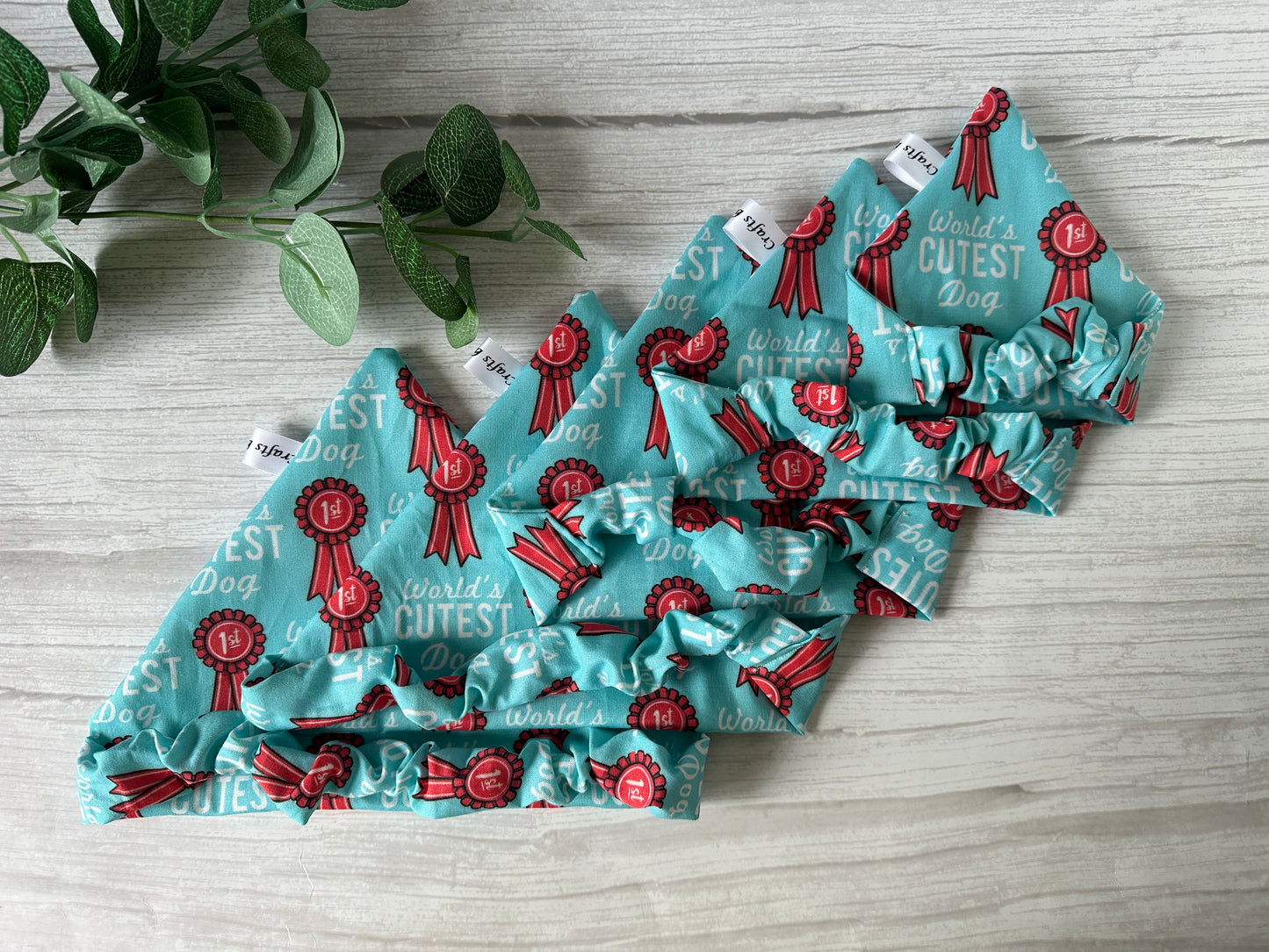 Four turquoise Cotton Pet Scrunchie Bandanas by Crafts by Kate, featuring red ribbons and "World’s Cutest Dog" print, are displayed on a light wooden surface beside a green leafy plant—a perfect handcrafted accessory for your furry friend.