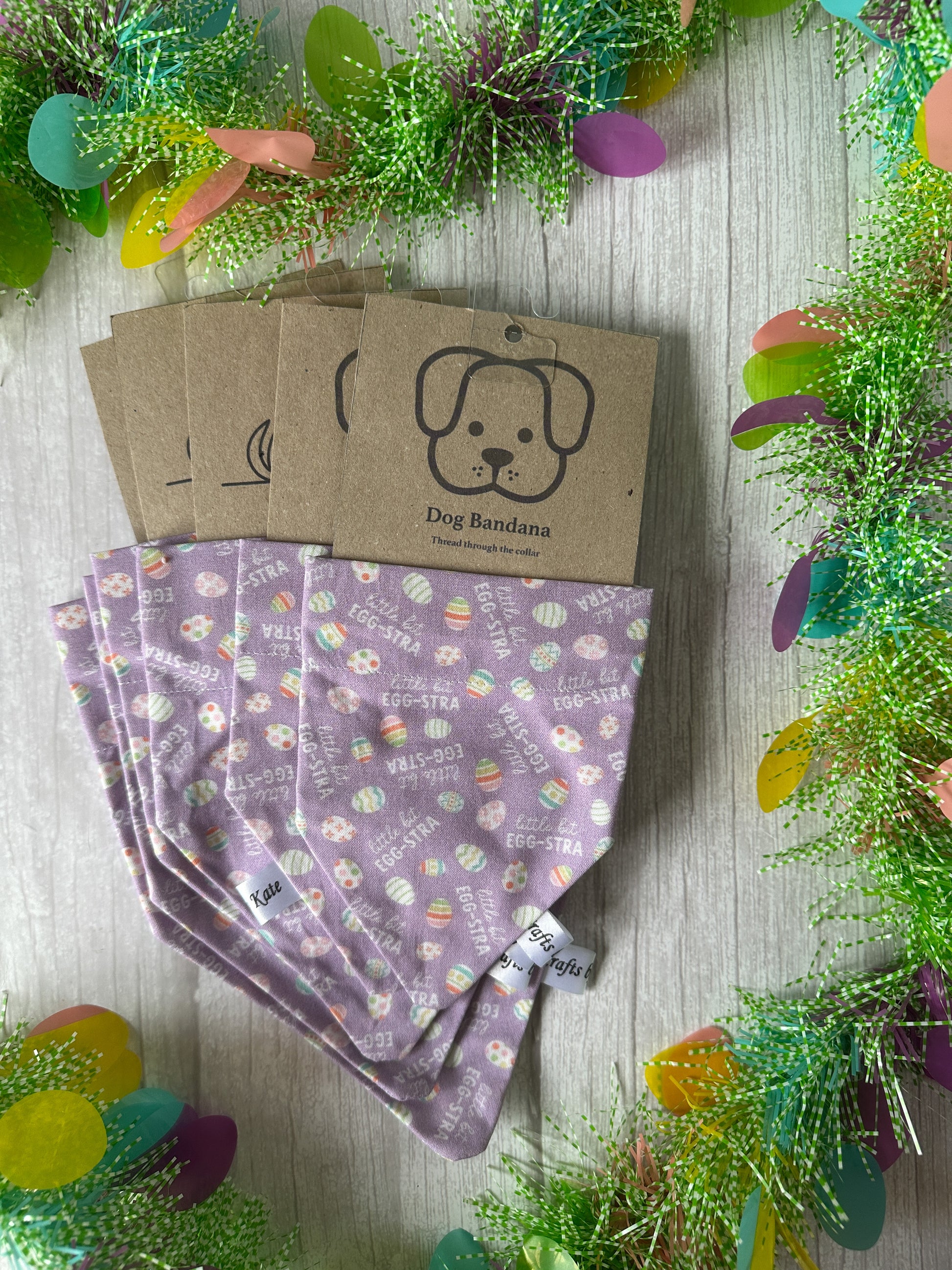Easter-themed purple dog bandanas from Crafts by Kate, named "Little bit Egg-stra," are displayed on a light wooden surface. Each handcrafted cotton bandana features a label with a cartoon dog's face and the words "Dog Bandana," surrounded by green, yellow, and purple decorative foliage.
