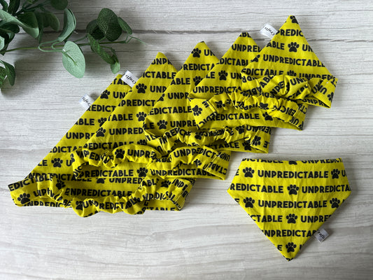 The Cotton Pet Scrunchie Bandana - Unpredictable by Crafts by Kate features a yellow cotton bandana with black "UNPREDICTABLE" text and paw prints, paired with matching scrunchies, displayed on light wood near green leaves.