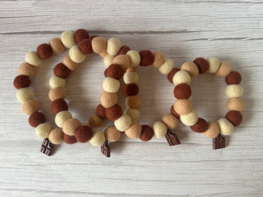 Three PomPom Pet Collars by Crafts by Kate, in cream, brown, and beige felt ball tones, are lined up on a light wooden surface. Perfect for chocolate lovers, each handcrafted piece boasts a small silver charm.