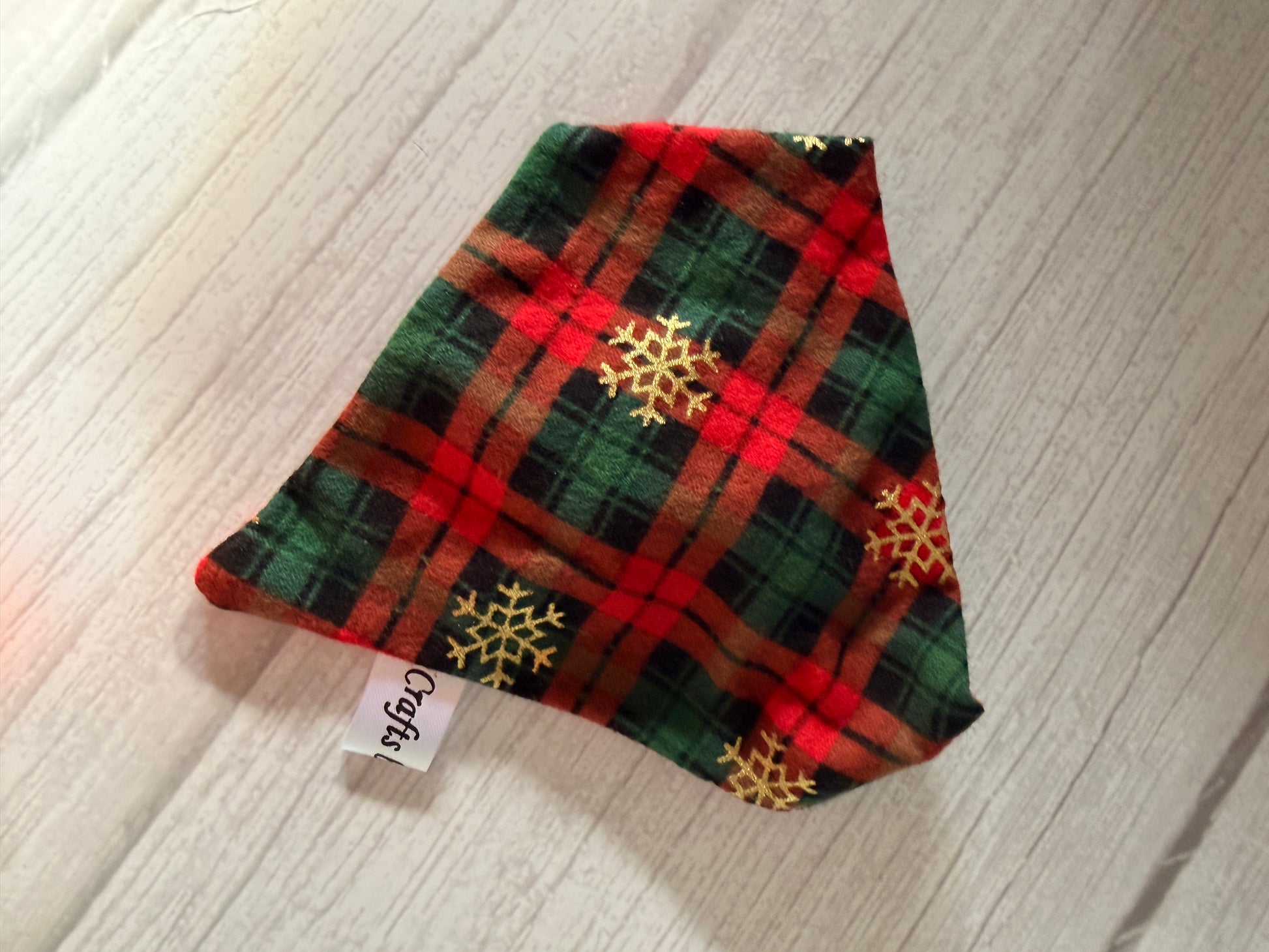 A red and green plaid triangle fabric with gold snowflakes and a "Crafty" tag rests on light wood. From Crafts by Kate, this Custom Order - AGL photography item is ideal as a tartan pet accessory or scrunchie bandana.