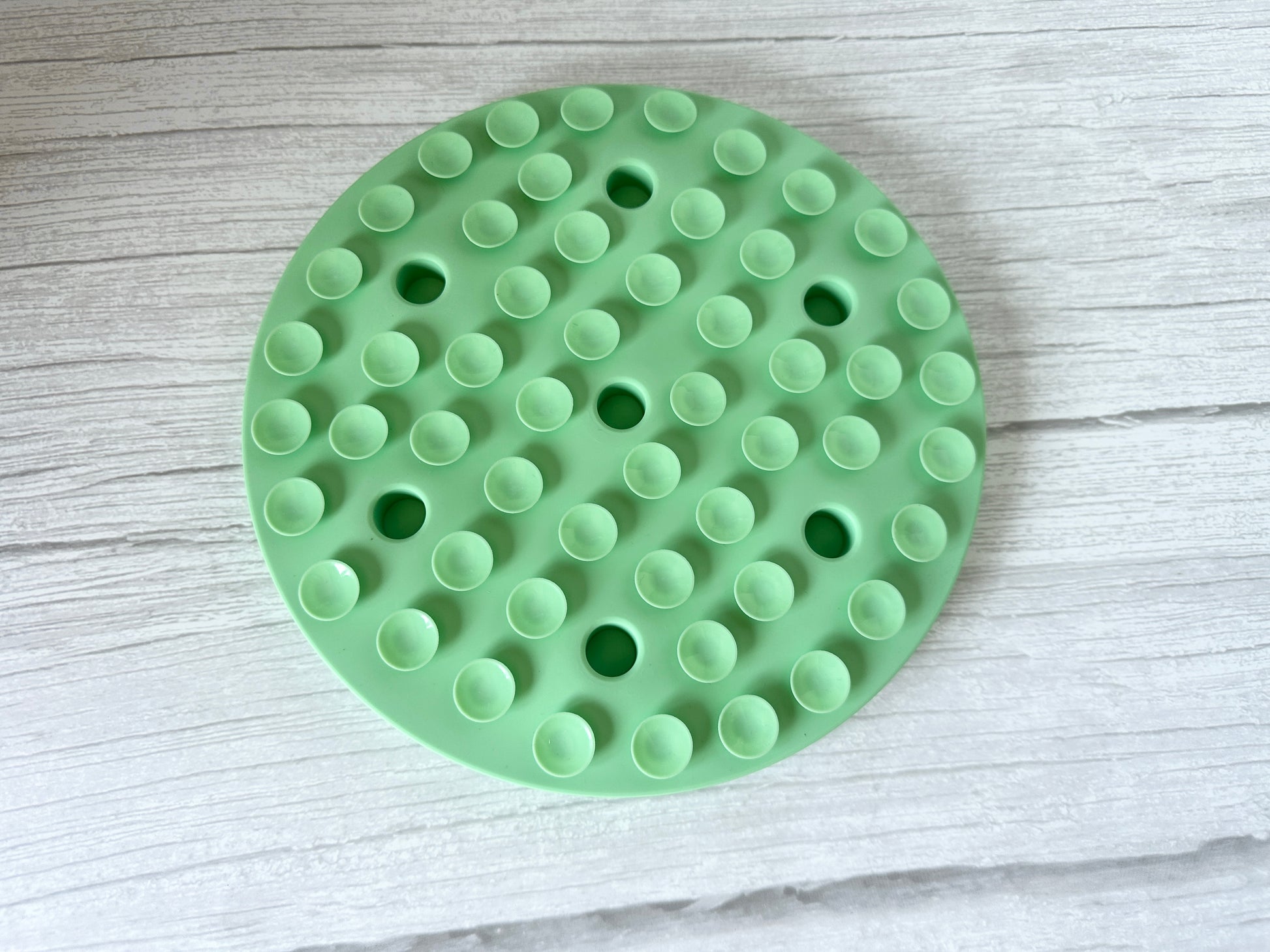 The Crafts by Kate Slow Feeder Pet Mat features a round, green silicone design with circular holes and discreet suction cups, placed on a light wooden surface.
