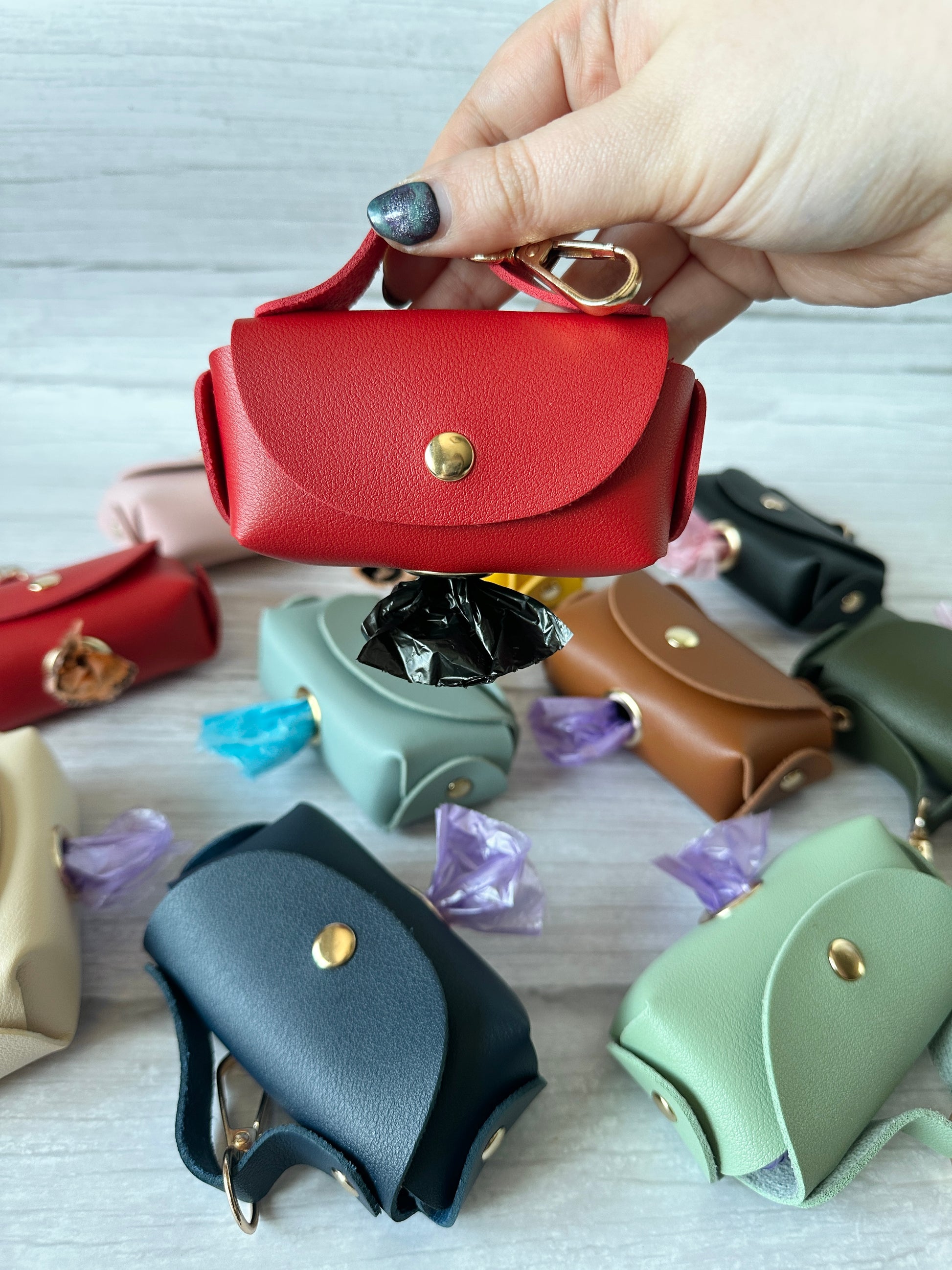 A hand holds a red Crafts by Kate Pet Poo Bag Holder Handbag in faux leather with a gold button and keyring, dispensing black bags from the bottom. Blue and green versions, embellished with decorative gems, are scattered on a wooden surface.