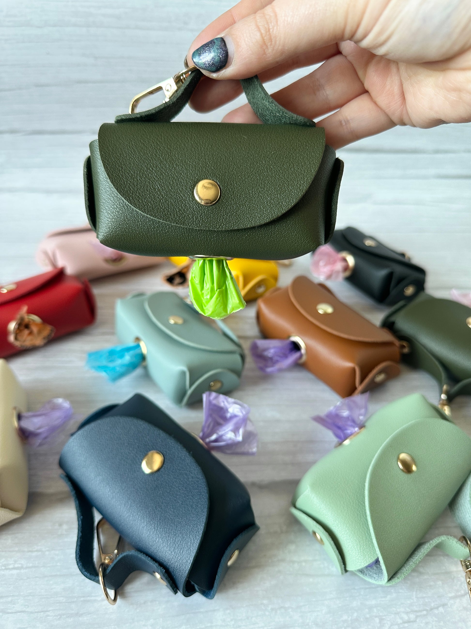A hand displays a small, handbag-shaped green leather pouch with a metal clasp, identified as Crafts by Kate's Pet Poo Bag Holder Handbag. In the background, various faux leather pouches in different colors rest on light wood, each showing a partially visible waste bag.