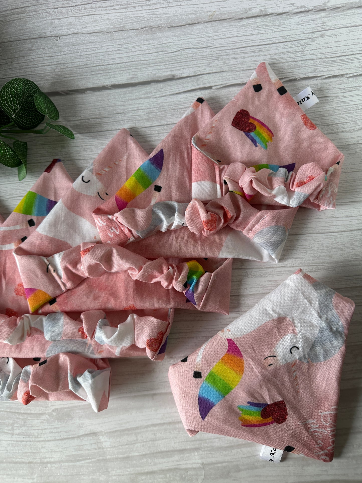 Five pink fabric headbands with rainbows, hearts, and smiling clouds are displayed on a white wood surface. In the top left corner is a small green potted plant next to Crafts by Kate's Cotton Pet Scrunchie Bandana - Bitch I’m a Unicorn.