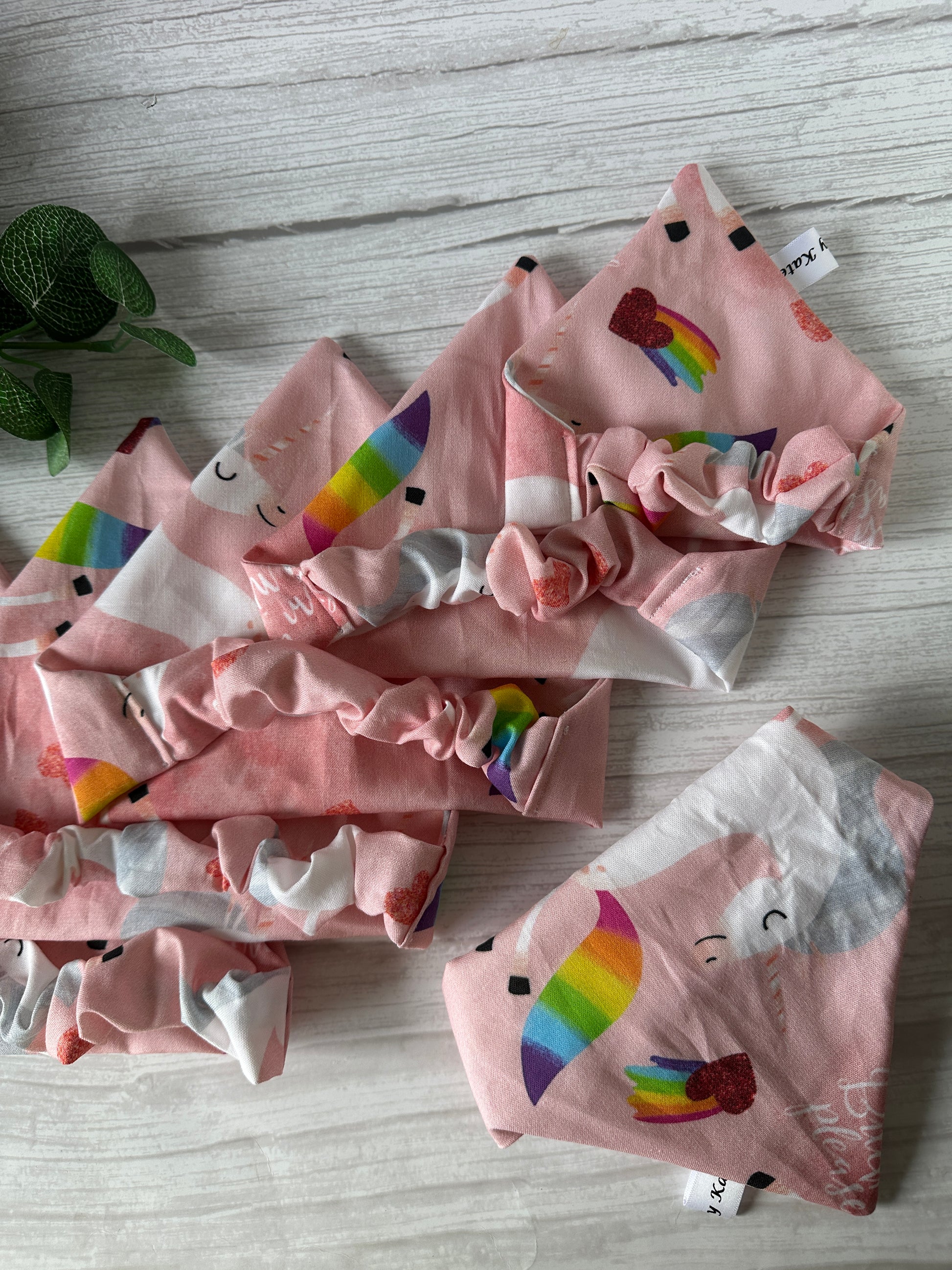 Five pink fabric headbands with rainbows, hearts, and smiling clouds are displayed on a white wood surface. In the top left corner is a small green potted plant next to Crafts by Kate's Cotton Pet Scrunchie Bandana - Bitch I’m a Unicorn.