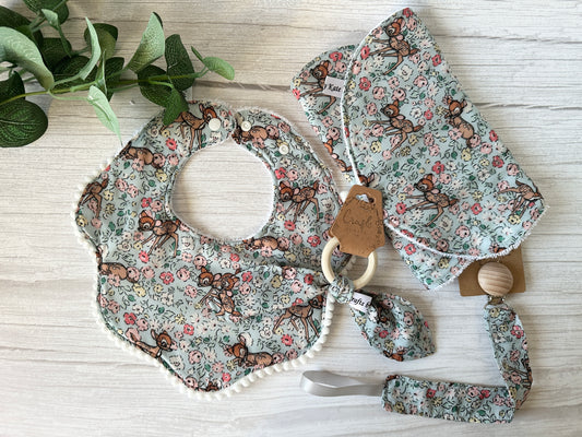 The Cotton & Terry Towelling Baby Gift Set - Floral Bambi by Crafts by Kate includes a soft cotton floral deer-patterned handcrafted bib, pacifier clip, and teether with a bamboo ring on light wood. A small green plant in the top left corner adds a natural touch.