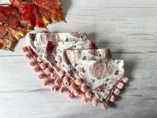 A collection of "Roses & Pumpkins" Cotton Pet Scrunchie Bandanas by Crafts by Kate is arranged in a fan shape on a light wooden surface. The bandanas feature pink pom-pom trim along the edges, with two orange autumn leaves placed in the top left corner, adding a touch of seasonal charm.