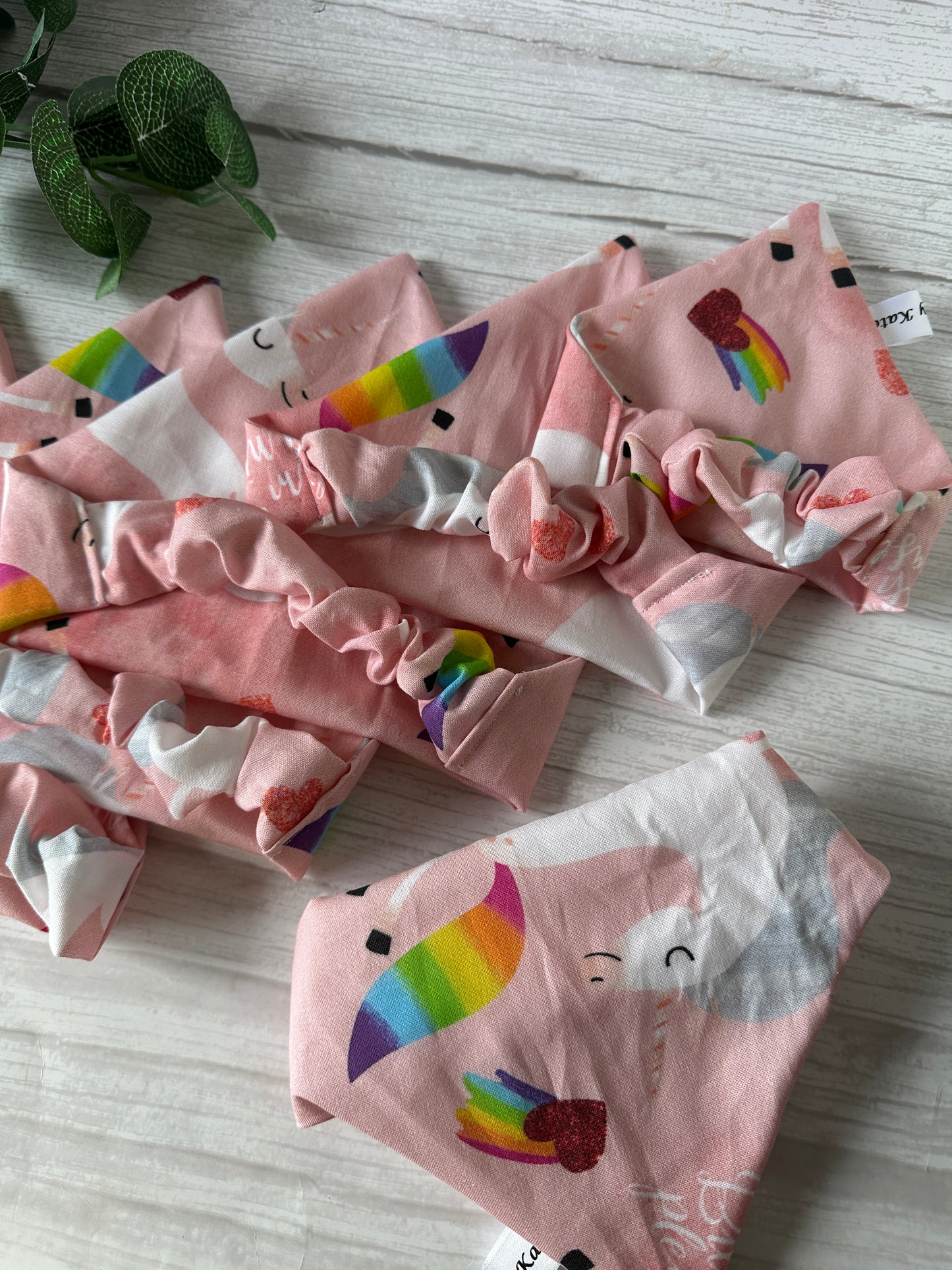 Several “Bitch I’m a Unicorn” Cotton Pet Scrunchie Bandanas by Crafts by Kate, featuring unicorn, rainbow, and heart patterns, are displayed on a white wooden surface next to a small green plant—a unique LGBTIQA+ pet accessory.