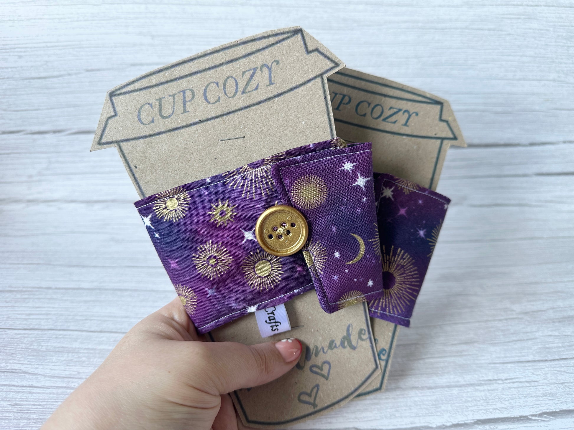 A hand holds two purple Cotton Reusable Cup Cozies - Midnight Sky by Crafts by Kate, showcasing gold celestial patterns with a button. Set against a wooden backdrop, the eco-friendly cozies have packaging labeled "Cup Cozy.