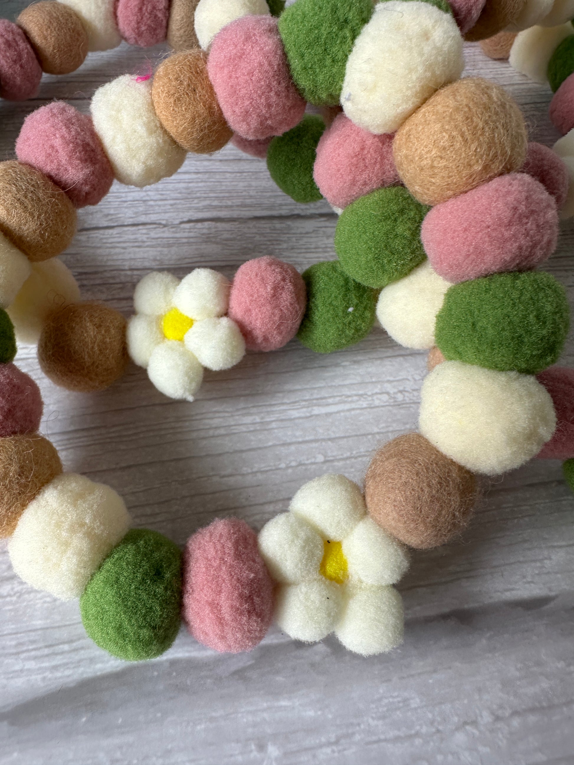 A close-up of the Crafts by Kate "PomPom Pet Collar - Fresh as a Daisy," featuring pink, white, green, and brown felt balls with small white daisy flowers and yellow centers, displayed on a light wooden surface.