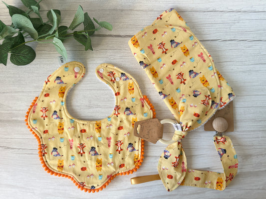 The "Cotton & Terry Towelling Baby Gift Set - Winnie the Pooh & Friends" by Crafts by Kate includes an ultra-soft bib and headband with charming cartoon animal prints like tigers and bunnies on light yellow fabric. The bib features orange trim and a wooden button, set against a light wooden background.