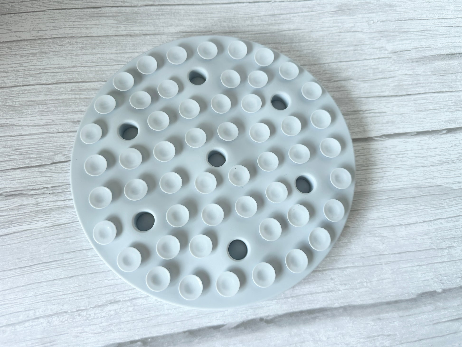 The Slow Feeder Pet Mat by Crafts by Kate is a round, white silicone mat with evenly spaced suction cups, set on a textured, light-colored wood surface. Its minimalist design features multiple small bumps that aid pet digestion and ensure easy cleaning.