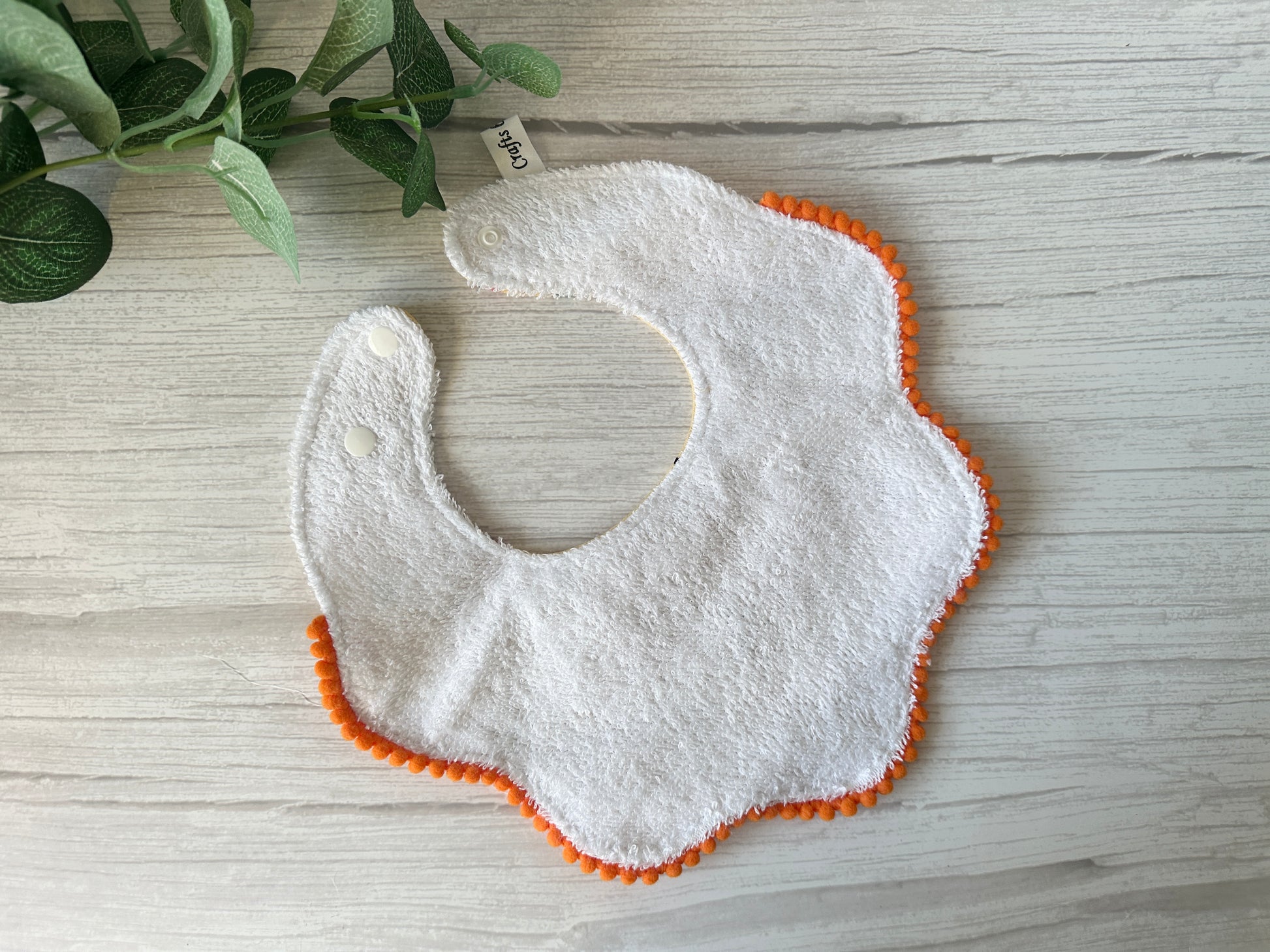 The Cotton & Terry Towelling Baby Gift Set - Winnie the Pooh & Friends by Crafts by Kate features a scalloped white baby bib with orange trim and snap buttons, set against a light wooden surface. A broad-leaved green plant is partially visible in the top left corner.