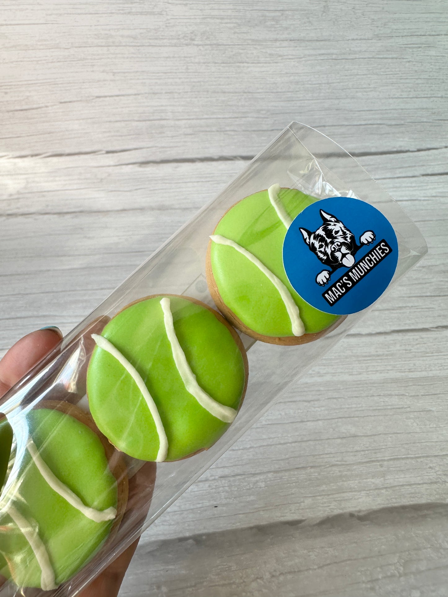 A hand holds a clear box with three tennis ball-shaped dog treats. A blue sticker shows a dog illustration and the Crafts by Kate brand. The label reads “Ball is Life - Arnie & Mac’s Goodies.”.