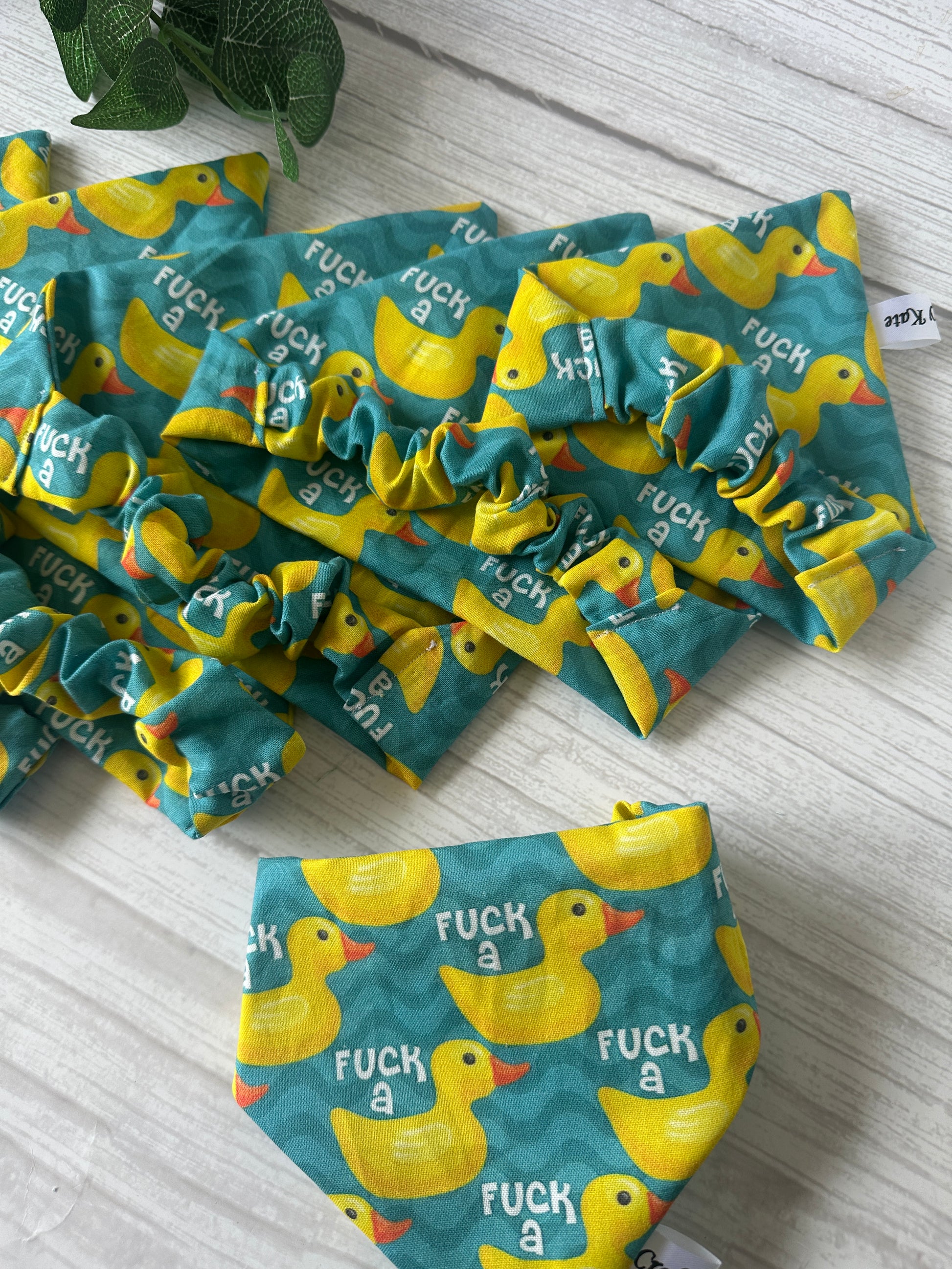 The Cotton Pet Scrunchie Bandana - F* a duck by Crafts by Kate features green and yellow fabric with yellow rubber duck prints and the phrase "F* a" repeated, displayed on a light wooden surface with leafy plant decor.