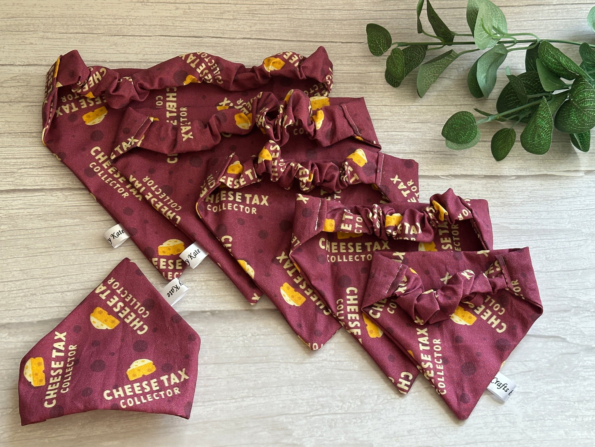 A selection of maroon handcrafted Cotton Pet Scrunchie Bandanas – Cheesetax Collector by Crafts by Kate, featuring cheese graphics, is displayed on a wooden surface. A small green plant in the top right corner adds a natural touch.