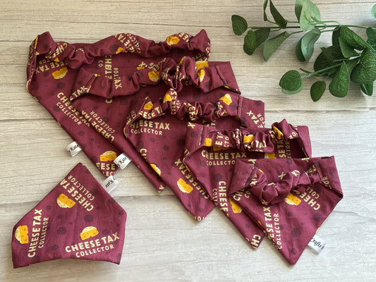 A selection of maroon handcrafted Cotton Pet Scrunchie Bandanas – Cheesetax Collector by Crafts by Kate, featuring cheese graphics, is displayed on a wooden surface. A small green plant in the top right corner adds a natural touch.