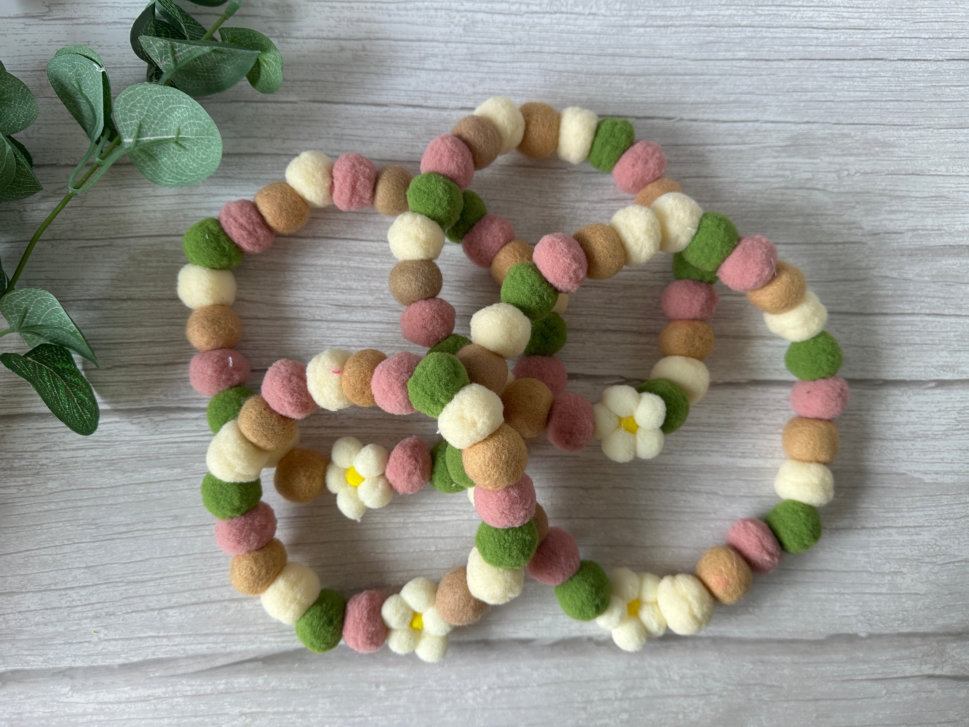 Three beaded garlands with soft felt balls in pink, green, beige, and cream—two featuring daisy flowers—are intertwined on light wood beside a plant, evoking the "PomPom Pet Collar - Fresh as a Daisy" by Crafts by Kate.