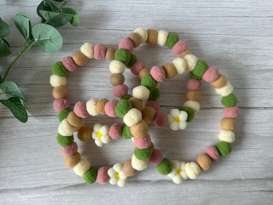Three beaded garlands with soft felt balls in pink, green, beige, and cream—two featuring daisy flowers—are intertwined on light wood beside a plant, evoking the "PomPom Pet Collar - Fresh as a Daisy" by Crafts by Kate.