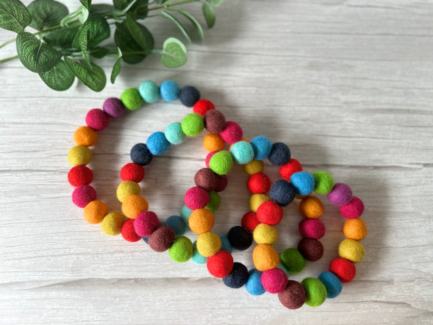 The PomPom Pet Collar - Rainbow by Crafts by Kate, adorned with vibrant shades, is beautifully displayed on a light wooden surface with a delicate sprig of green leaves in the background.