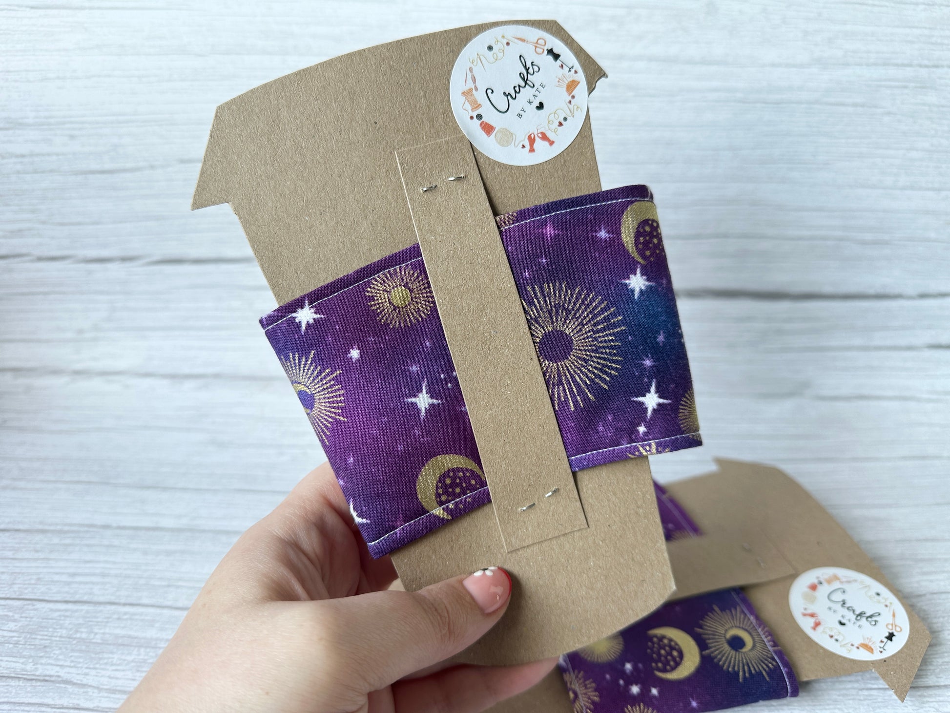A hand holds a coffee cup wrapped in the "Cotton Reusable Cup Cozy - Midnight Sky" by Crafts by Kate, showcasing celestial designs in purple, gold, and white with stars and suns. A “Create” sticker adds handcrafted charm to the cozy.