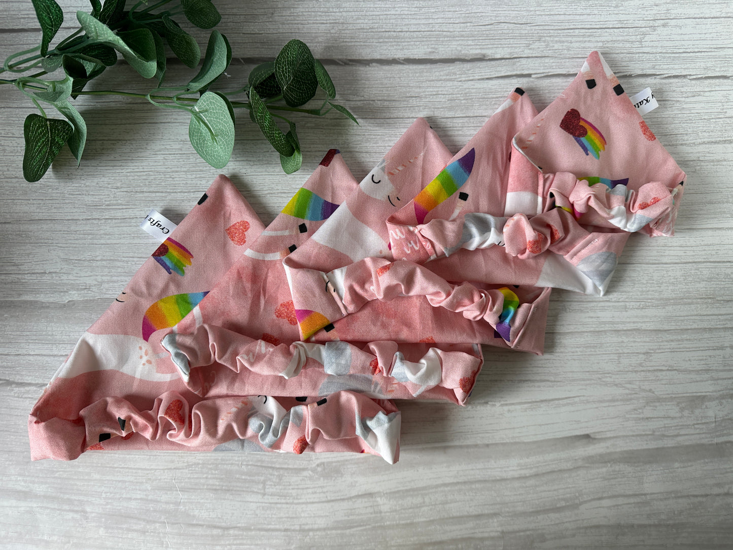 Four pink Cotton Pet Scrunchie Bandanas—“Bitch I’m a Unicorn” by Crafts by Kate—featuring rainbow, heart, and smiley face patterns, are displayed on light wood beside a leafy plant—a cheerful LGBTIQA+ pet accessory.