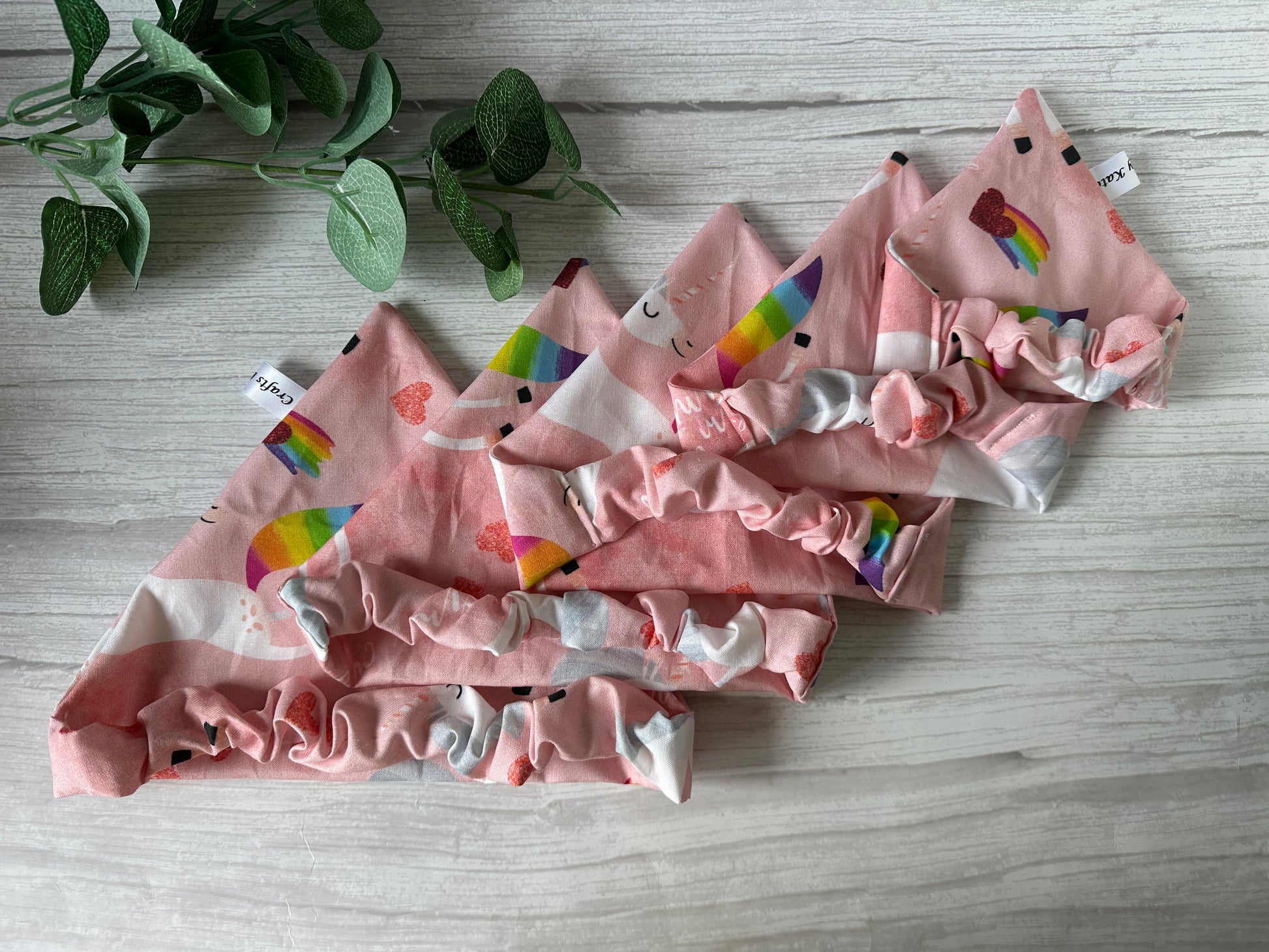Four pink Cotton Pet Scrunchie Bandanas—“Bitch I’m a Unicorn” by Crafts by Kate—featuring rainbow, heart, and smiley face patterns, are displayed on light wood beside a leafy plant—a cheerful LGBTIQA+ pet accessory.