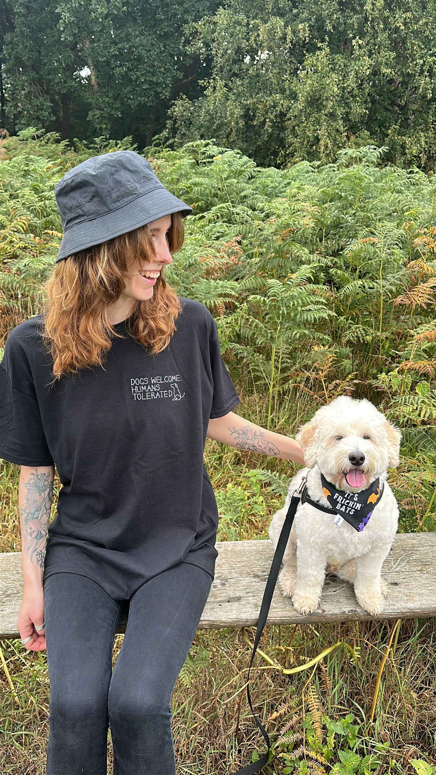 A woman in a Crafts by Kate "Dogs welcome" oversized cotton t-shirt and black hat sits on an outdoor bench, smiling at her fluffy white dog in a harness, surrounded by lush ferns and trees.