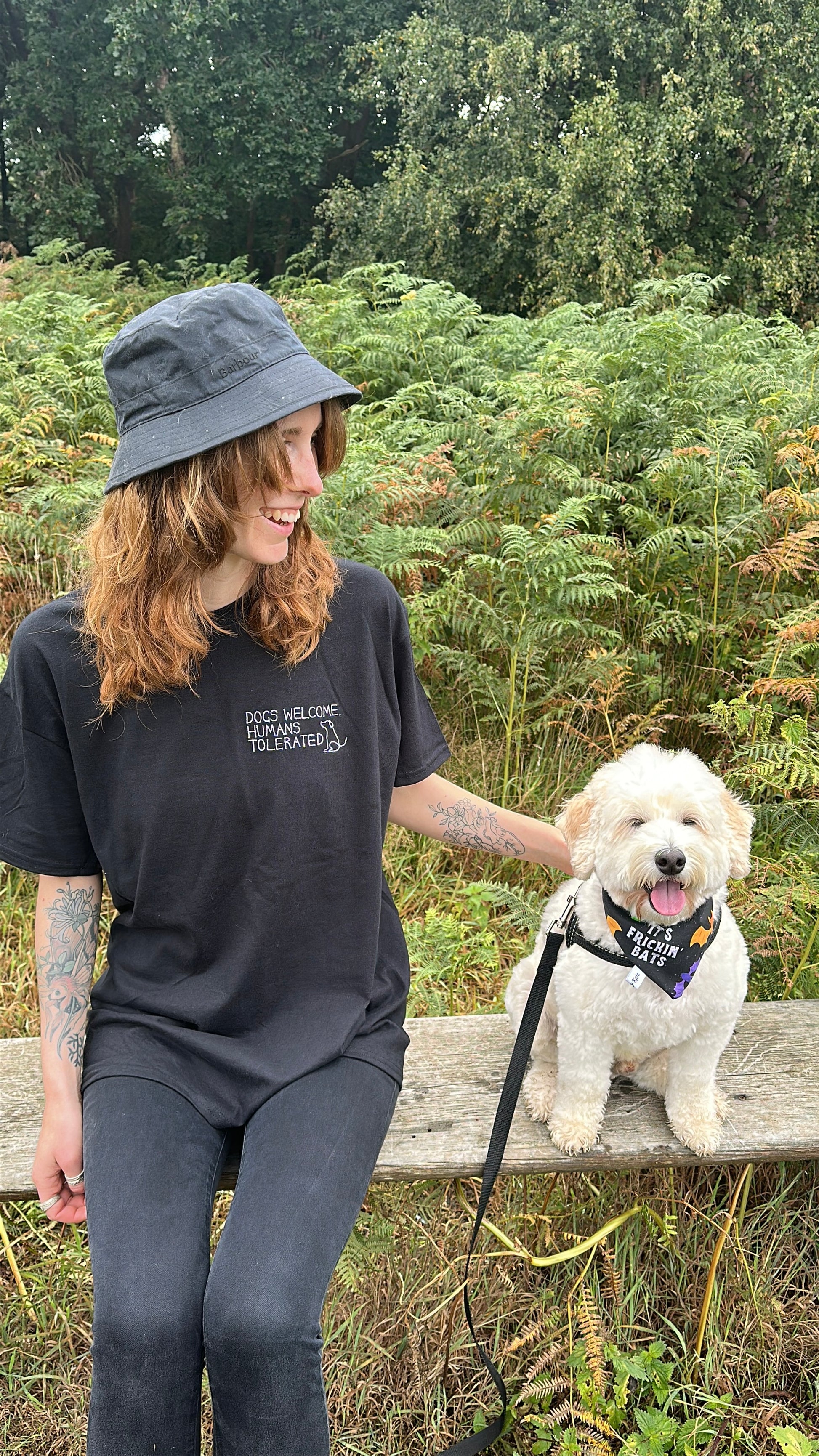 A woman in a Crafts by Kate "Dogs welcome" oversized cotton t-shirt and black hat sits on an outdoor bench, smiling at her fluffy white dog in a harness, surrounded by lush ferns and trees.