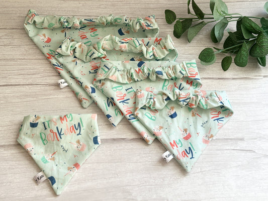 Crafts by Kate's Cotton Pet Scrunchie Bandanas, featuring colorful "It’s My Barkday" text and playful dog illustrations, lay on a light wooden surface with a green leafy plant in the top right corner. Handcrafted tags labeled "XSmall" are visible.