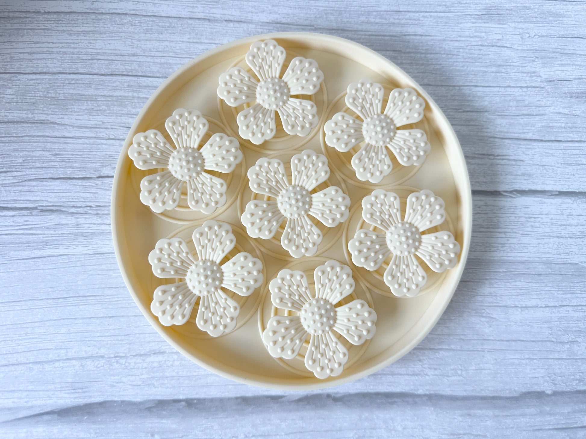 The Crafts by Kate Slow Feeder Pet Mat showcases a round tray with seven white flower-shaped cookies on a textured light wooden surface. It offers mess-free serving, featuring washable materials and secure suction cups to keep it in place.