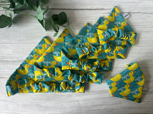 A green and yellow Cotton Pet Scrunchie Bandana—F*** a duck by Crafts by Kate—featuring a rubber duck print, displayed on light wood beside a green plant.