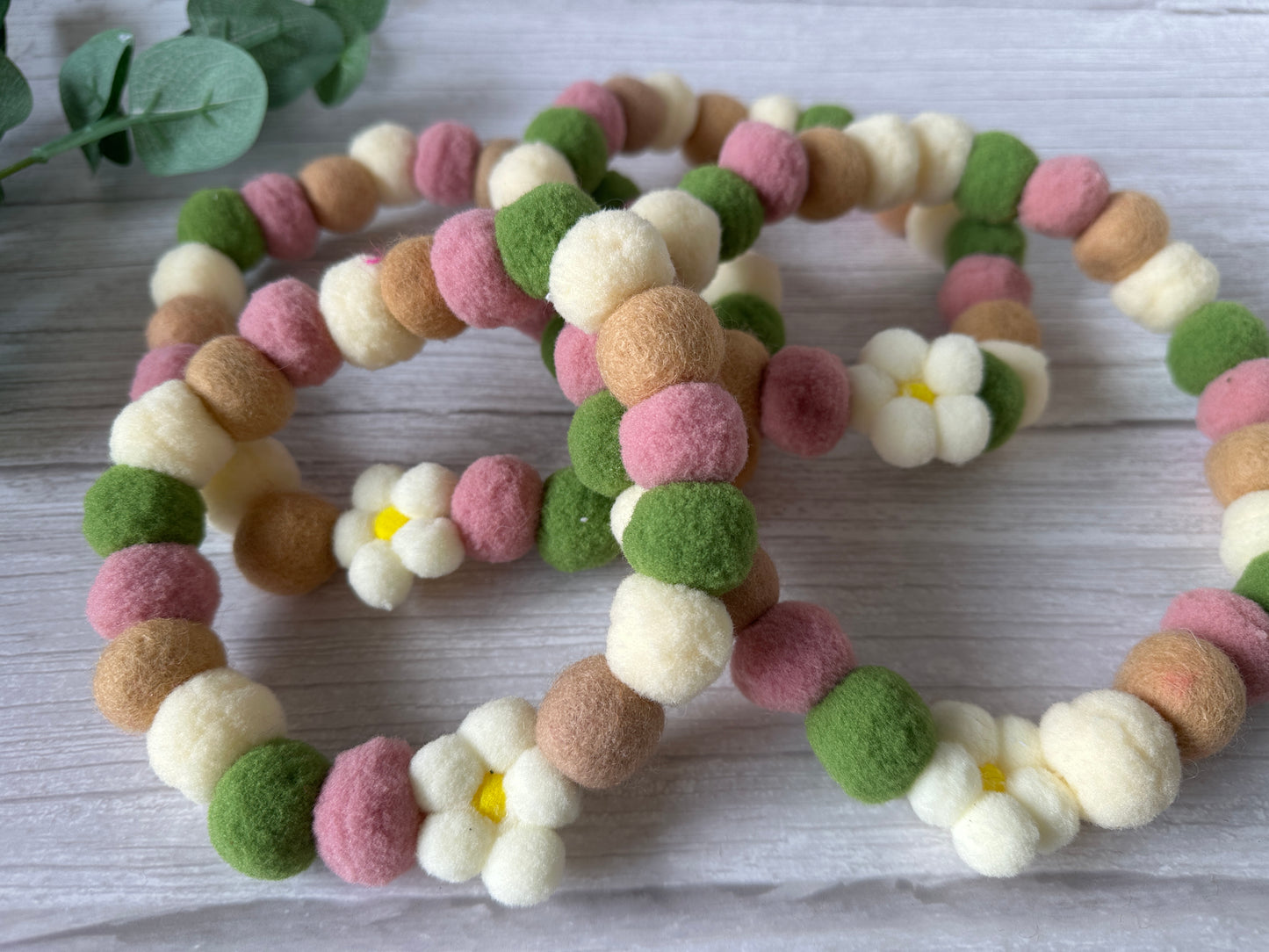 A close-up of the PomPom Pet Collar - Fresh as a Daisy by Crafts by Kate, featuring pink, green, brown, and cream beads with daisy-shaped accents, displayed on a light wooden surface.
