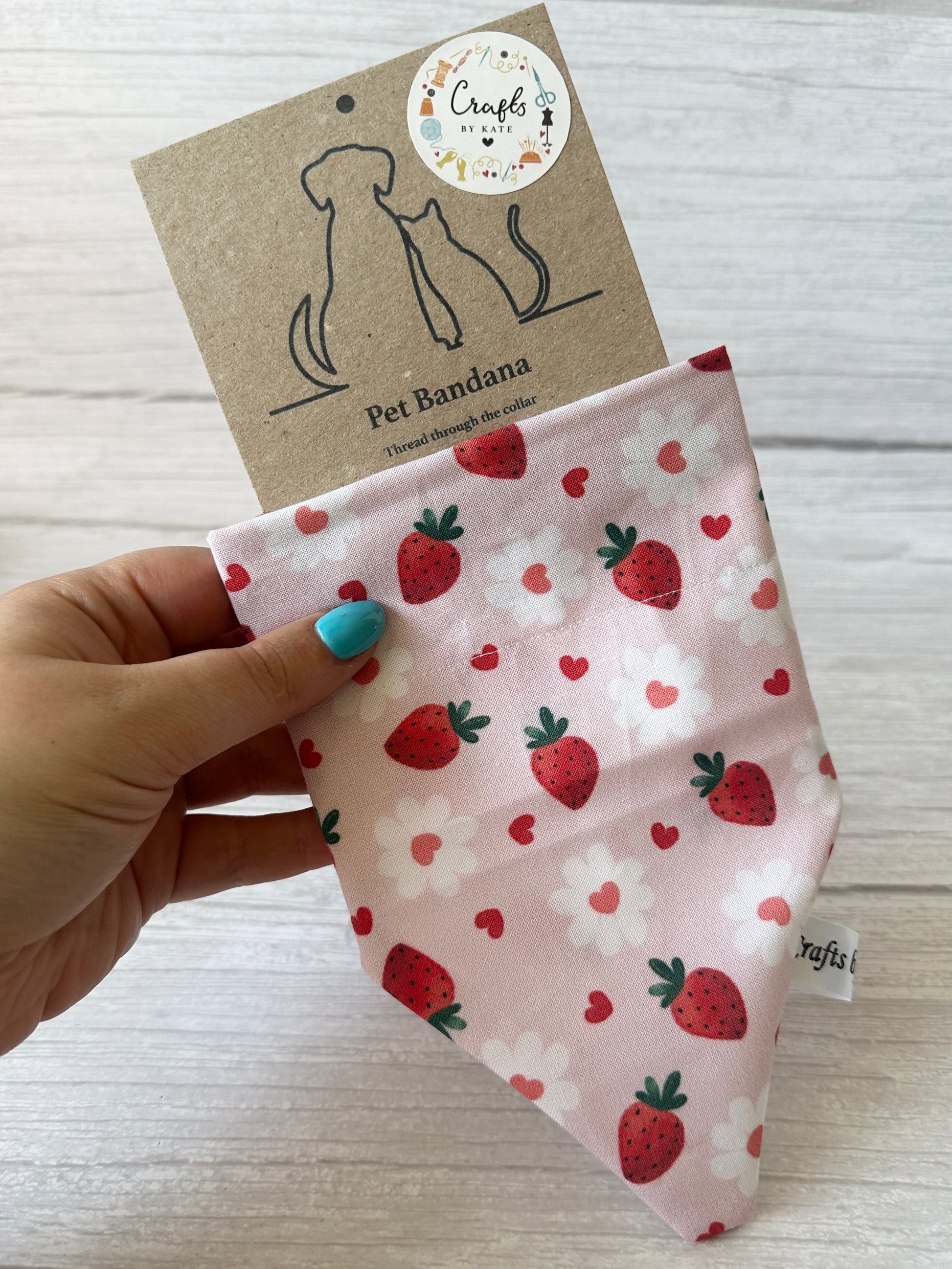 A hand holds the Strawberries & Daisies pet bandana by Crafts by Kate. Decorated with strawberries, daisies, and hearts, it's tagged "Arnie & Mac’s Goodies"—a playful accessory for pets who love fun and treats.