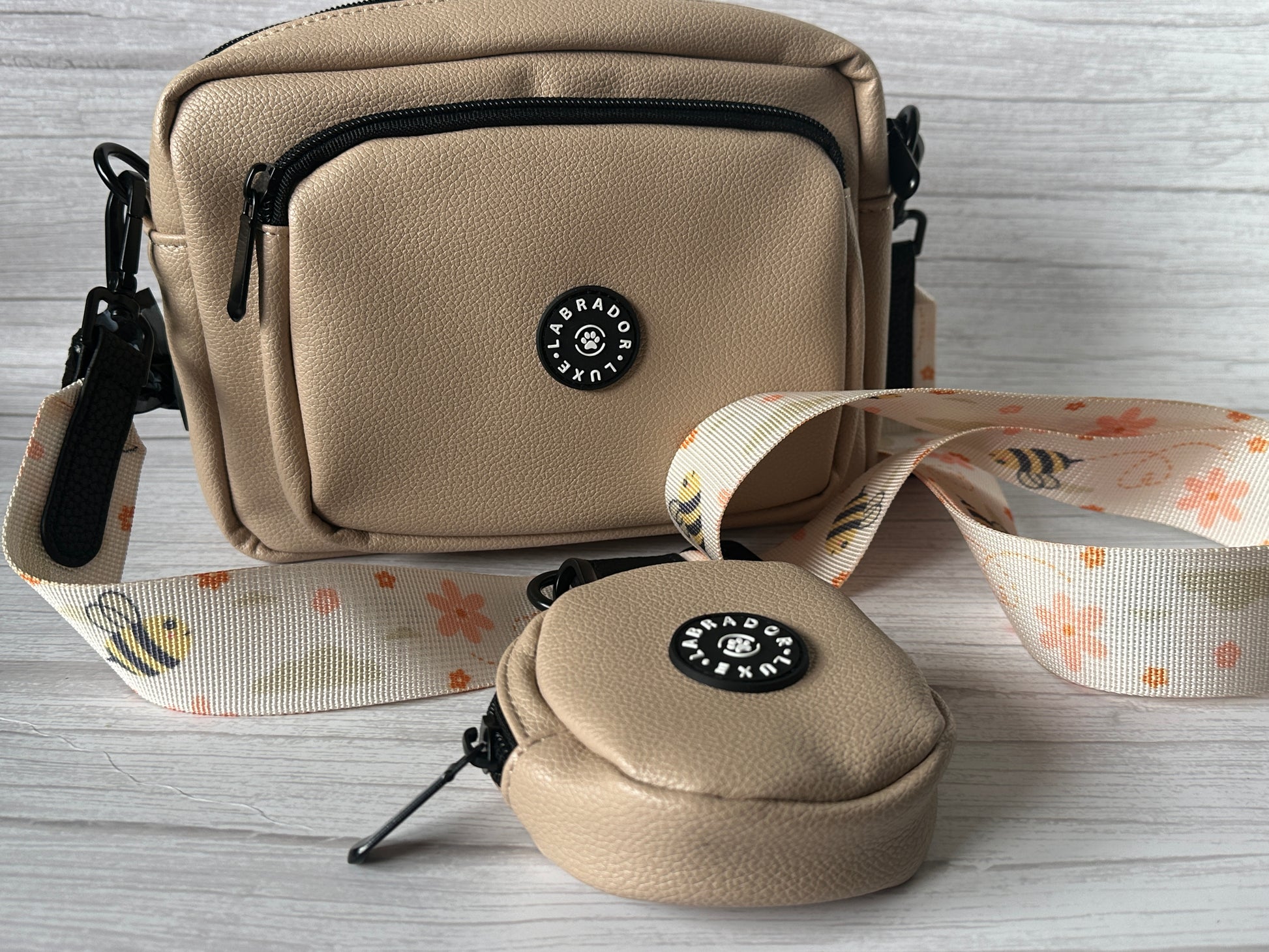 The Bumble Dog Walking Bag by Crafts by Kate is a beige bag with a matching round coin pouch, black zippers, and a floral bee strap, shown on a light wooden surface.