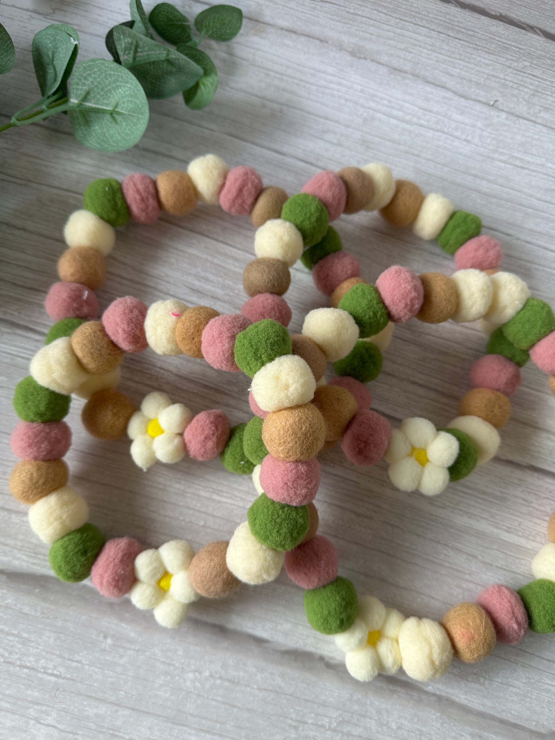 Five PomPom Pet Collar - Fresh as a Daisy garlands by Crafts by Kate, featuring soft cream, pink, green, and brown felt balls with white daisy flowers and yellow centers, are arranged on light wood beside green leaves.