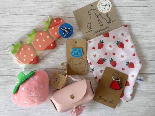 A flat lay of Crafts by Kate’s Strawberries & Daisies - Arnie & Mac’s Goodies, featuring a strawberry-print bandana, strawberry toys, a pink plush strawberry, waste bag holder, and keychain on light wood.