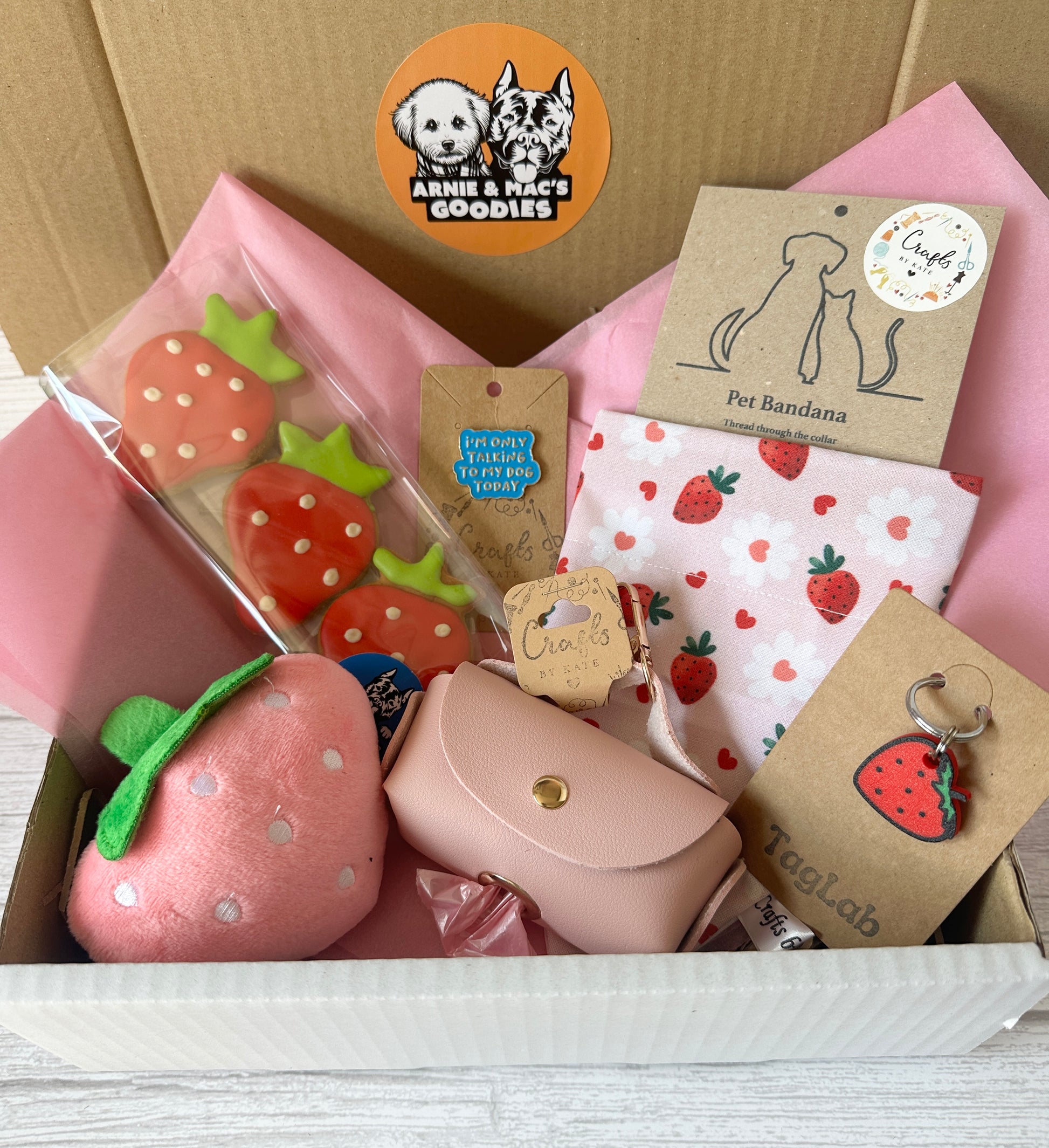 The Strawberries & Daisies - Arnie & Mac’s Goodies gift box by Crafts by Kate includes strawberry dog treats, a pink plush toy, waste bag holder, strawberry bandana, and a cute keychain—beautifully wrapped in pink tissue paper.