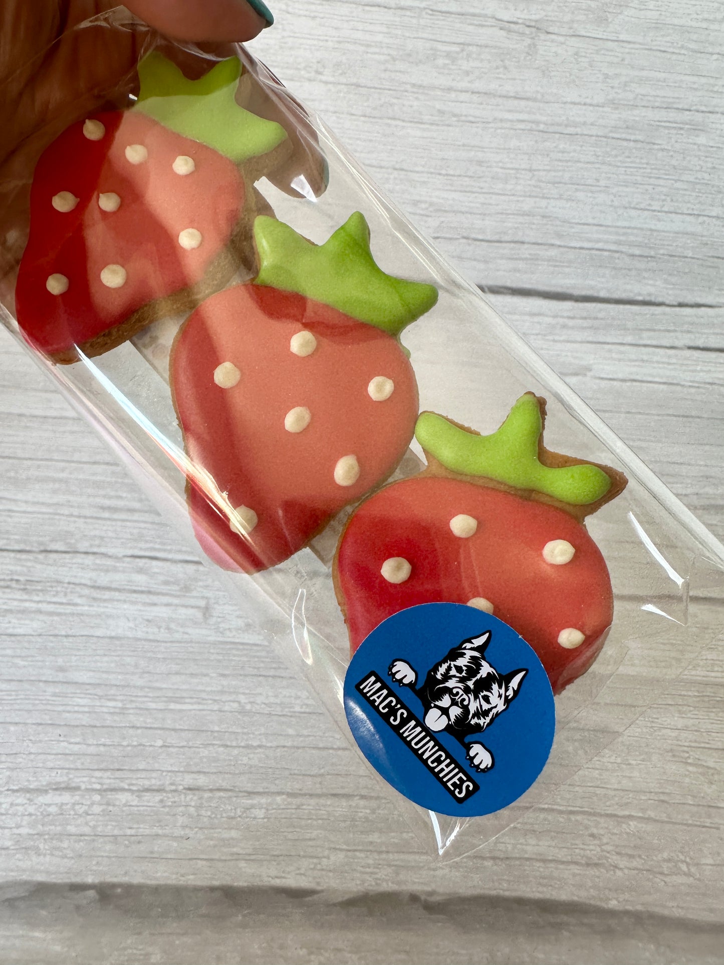 A hand holds a clear bag with three strawberry-shaped cookies decorated with red icing and white dots—Strawberries & Daisies by Crafts by Kate, from Arnie & Mac’s Goodies, as shown on the blue sticker featuring a dog illustration.