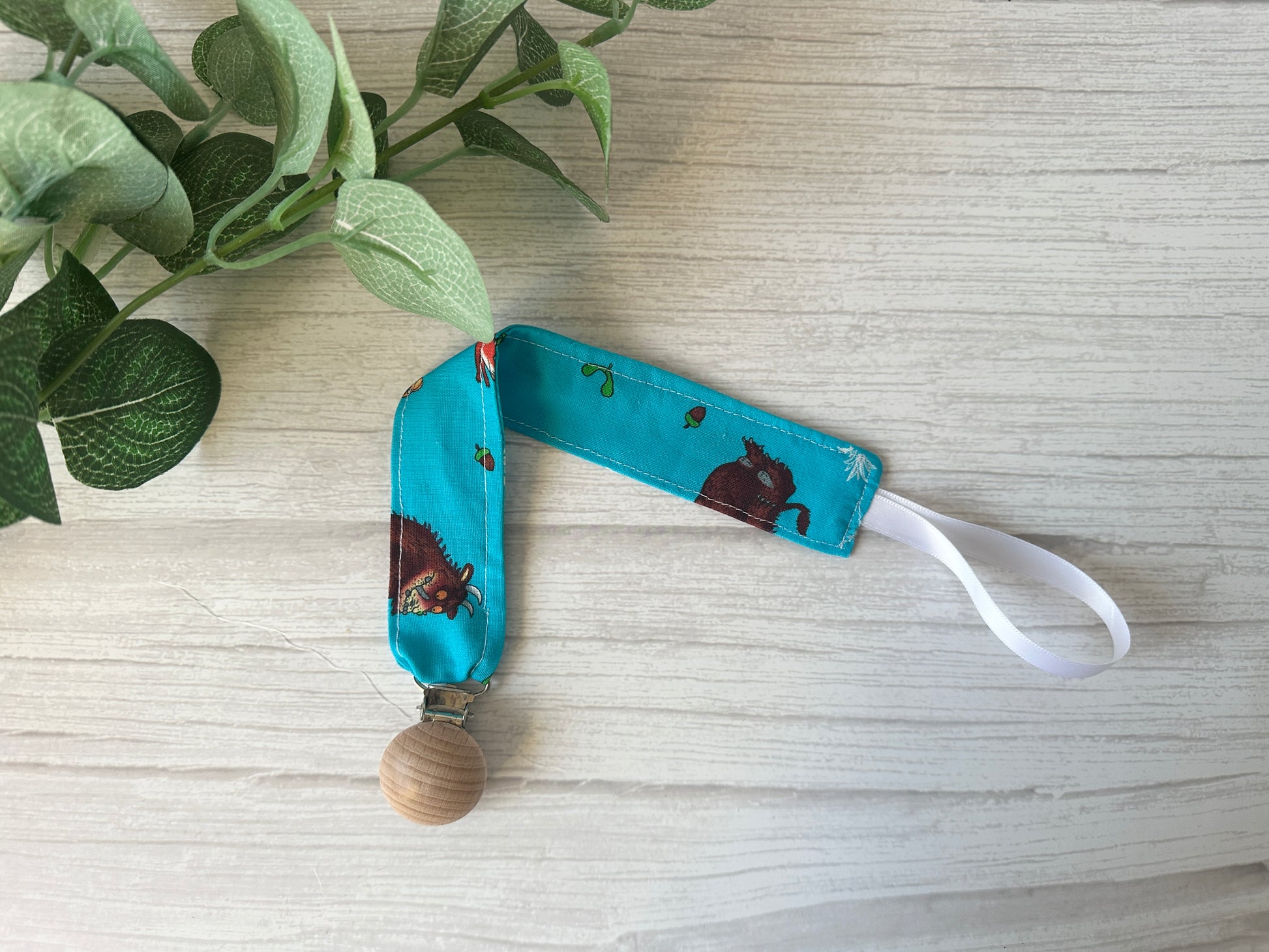 The Crafts by Kate Cotton & Terry Towelling Baby Gift Set - Gruffalo in blue, featuring a pacifier clip with a wooden clasp and ultra-soft cotton in turquoise with cartoon animal prints, is displayed on light wood next to green leaves—an ideal addition to handcrafted baby accessories.