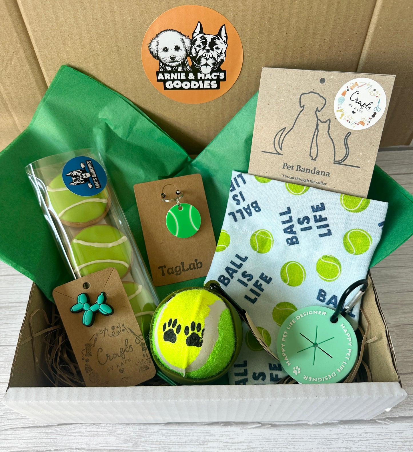The "Ball is Life - Arnie & Mac’s Goodies" gift box by Crafts by Kate includes a tennis ball toy, dog treats, blue paw keychain, tennis ball pet tag, pet bandana, and round tag—beautifully packed in green tissue with a cute dog sticker.